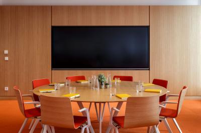 Salle de réunion avec table ronde en bois, chaises rouges, écran mural noir et fournitures de bureau disposées.
