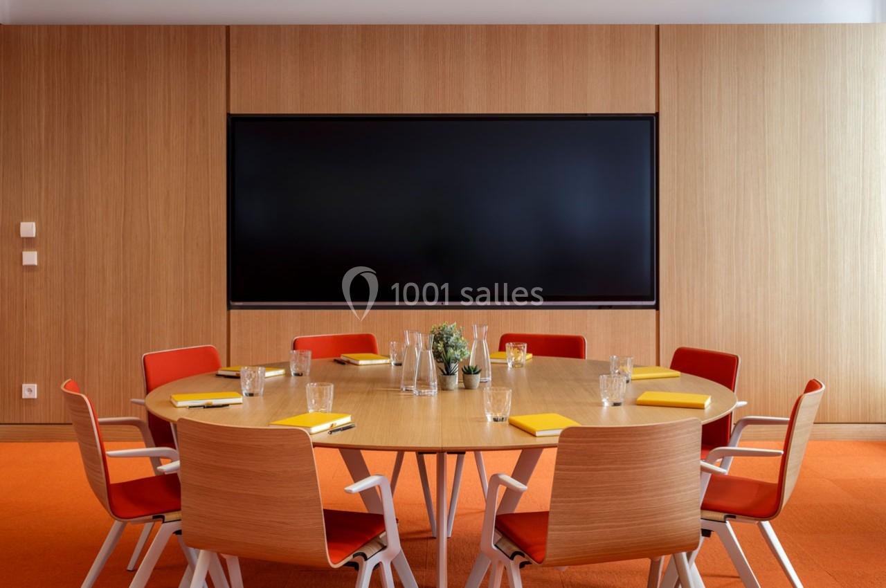 Salle de réunion avec table ronde en bois, chaises rouges, écran mural noir et fournitures de bureau disposées.