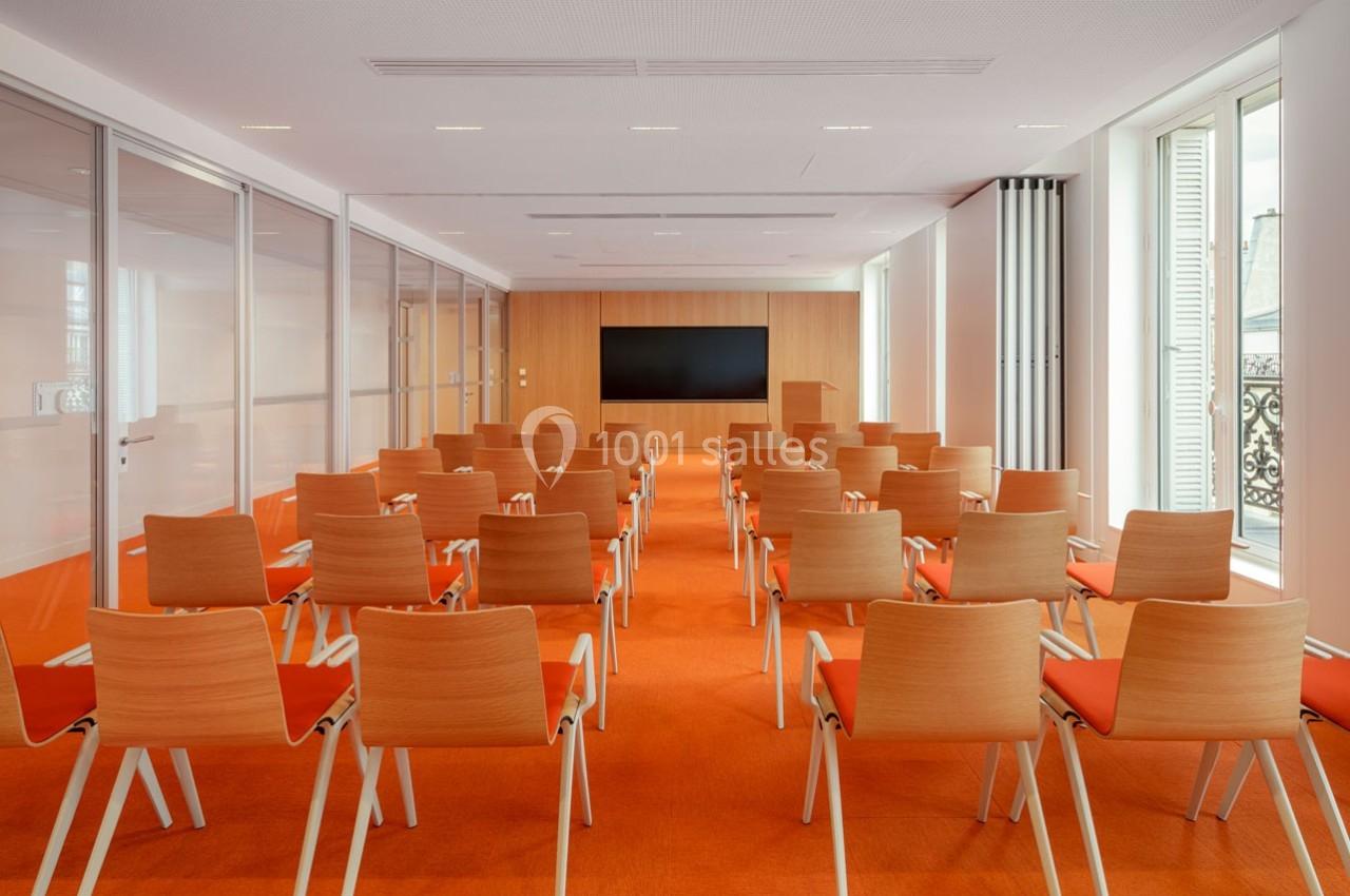 Salle de conférence lumineuse avec des rangées de chaises en bois et un écran noir fixé sur un mur en bois clair.