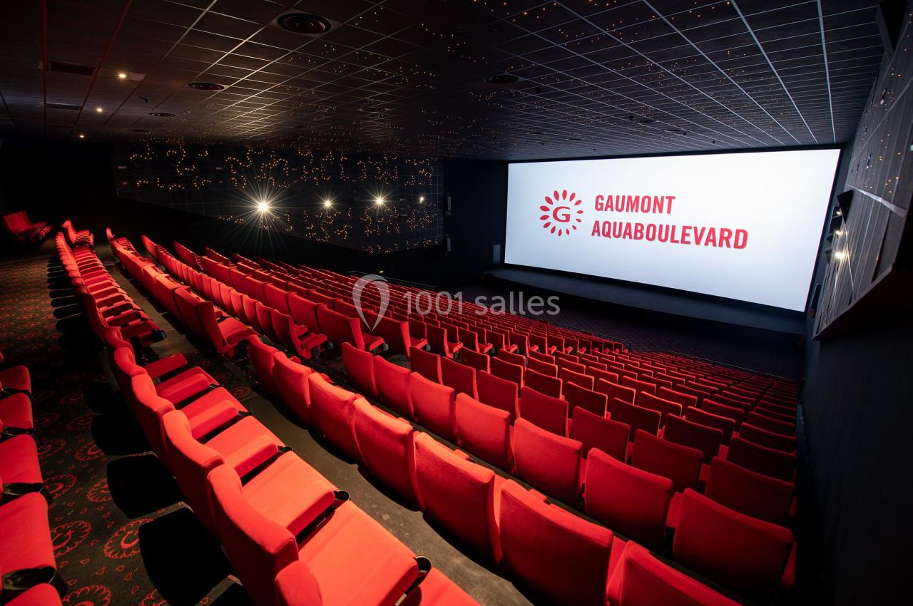 Salle de cinéma avec rangées de sièges rouges vides, grand écran affichant ’Gaumont Aquaboulevard’.