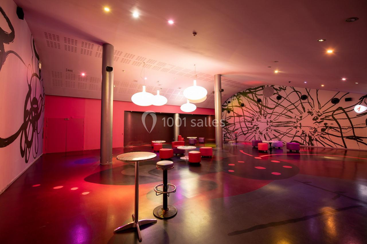 Salle moderne avec éclairage coloré, murs décorés de motifs abstraits, tables hautes et sièges dispersés.