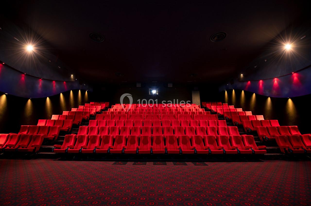 Salle de cinéma vide avec des rangées de sièges rouges et un écran blanc au fond, éclairée par des lumières tamisées.