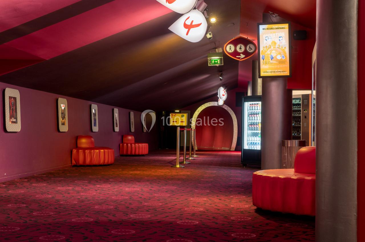 Couloir d'un cinéma avec moquette rouge, sièges arrondis, distributeurs automatiques et signalétique lumineuse.