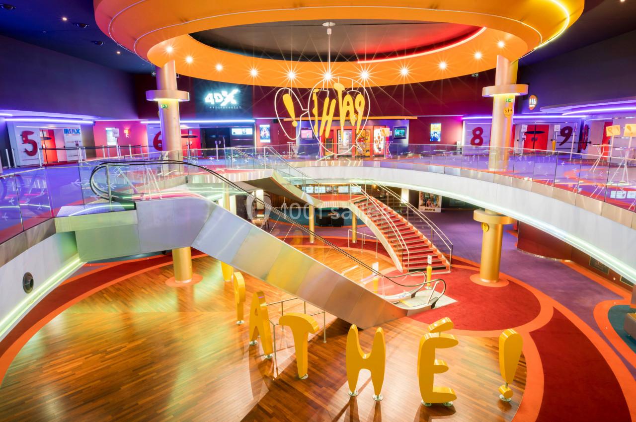 Intérieur lumineux d'un cinéma avec escalators, escalier central, enseigne ’Pathé’ et ambiance colorée.