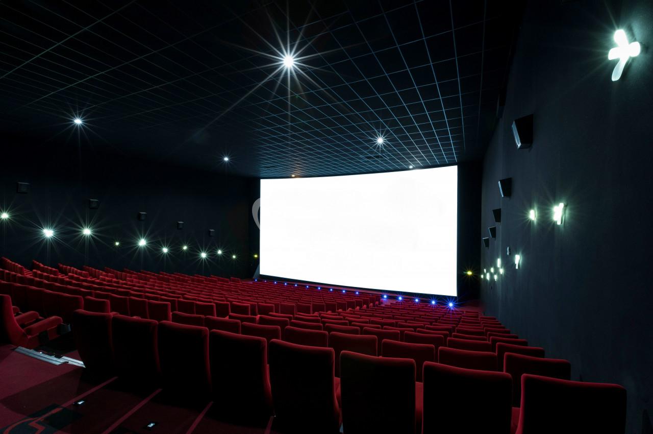 Salle de cinéma vide avec rangées de sièges rouges et un grand écran blanc allumé sous un plafond sombre.