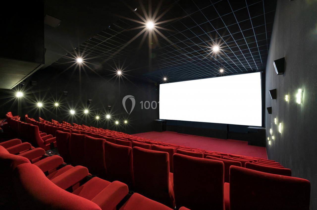 Salle de cinéma vide avec des rangées de fauteuils rouges, un écran blanc et un éclairage tamisé.