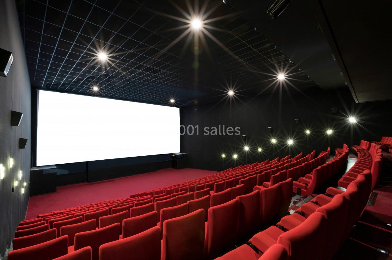 Salle de cinéma vide avec des rangées de sièges rouges, un écran blanc allumé et un plafond sombre éclairé.