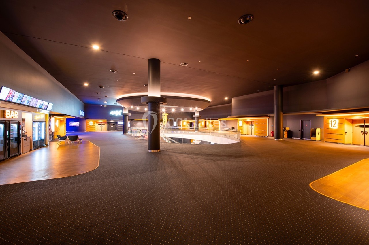 Hall spacieux d'un cinéma avec moquette sombre, éclairage tamisé, bar et accès aux salles numérotées.