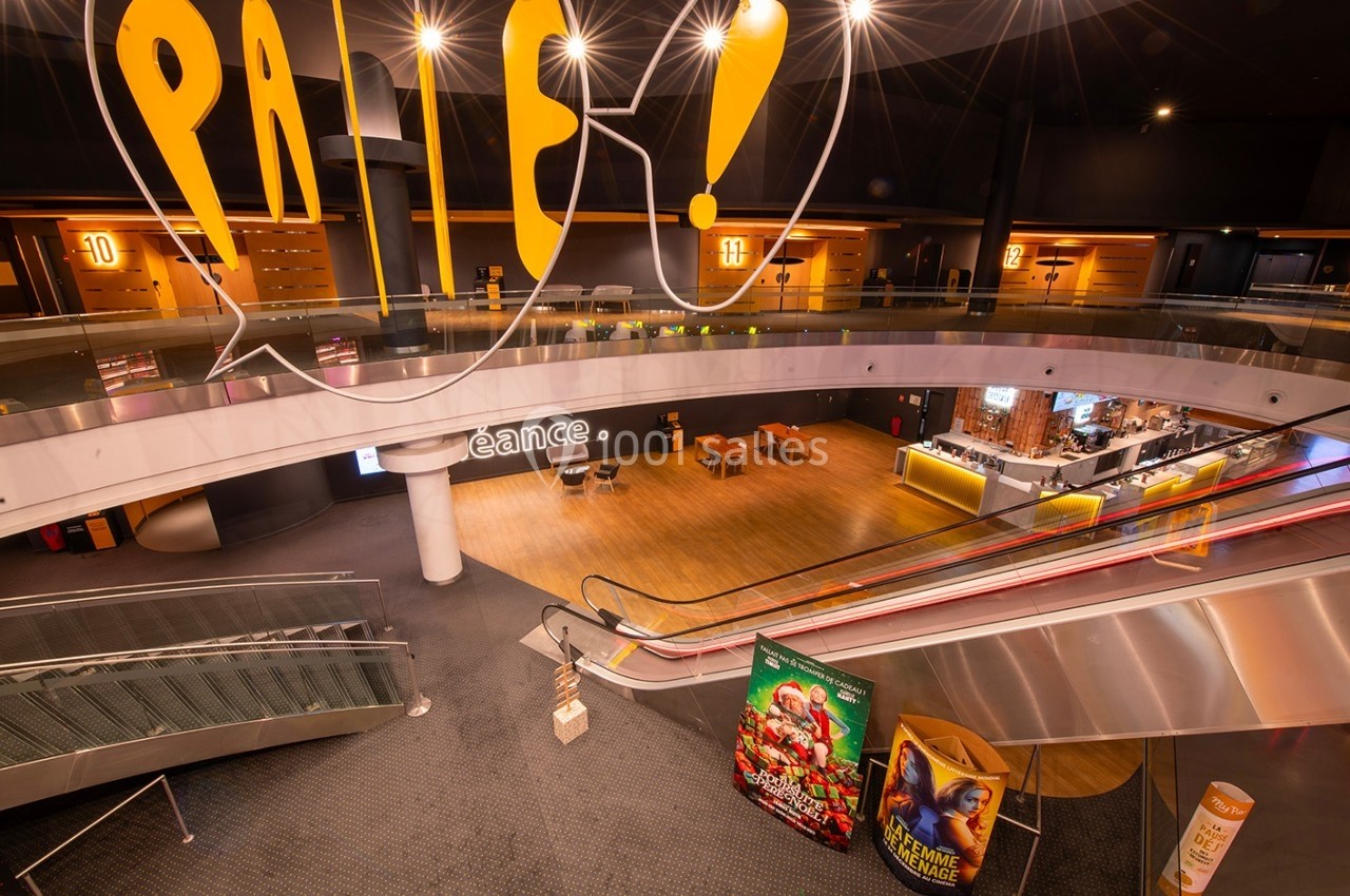 Vue intérieure d'un cinéma avec escalators, affiches de films, comptoir de confiserie et signalétique lumineuse.