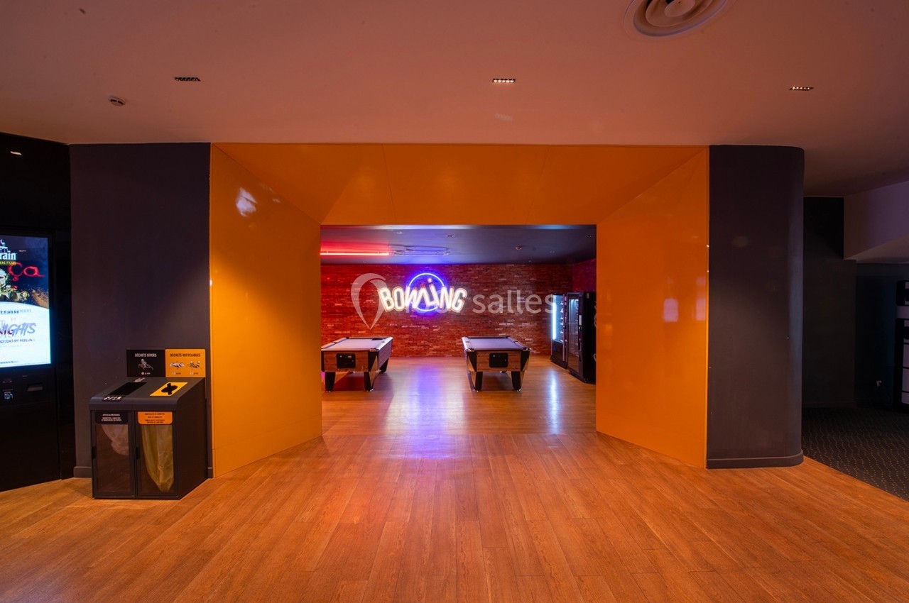 Entrée d'une salle de bowling avec un mur en briques, un néon ’Bowling’ et des tables de jeu en premier plan.