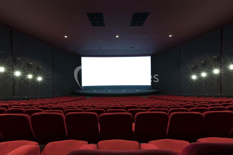 Salle de cinéma vide avec des rangées de sièges rouges et un écran blanc allumé au fond.