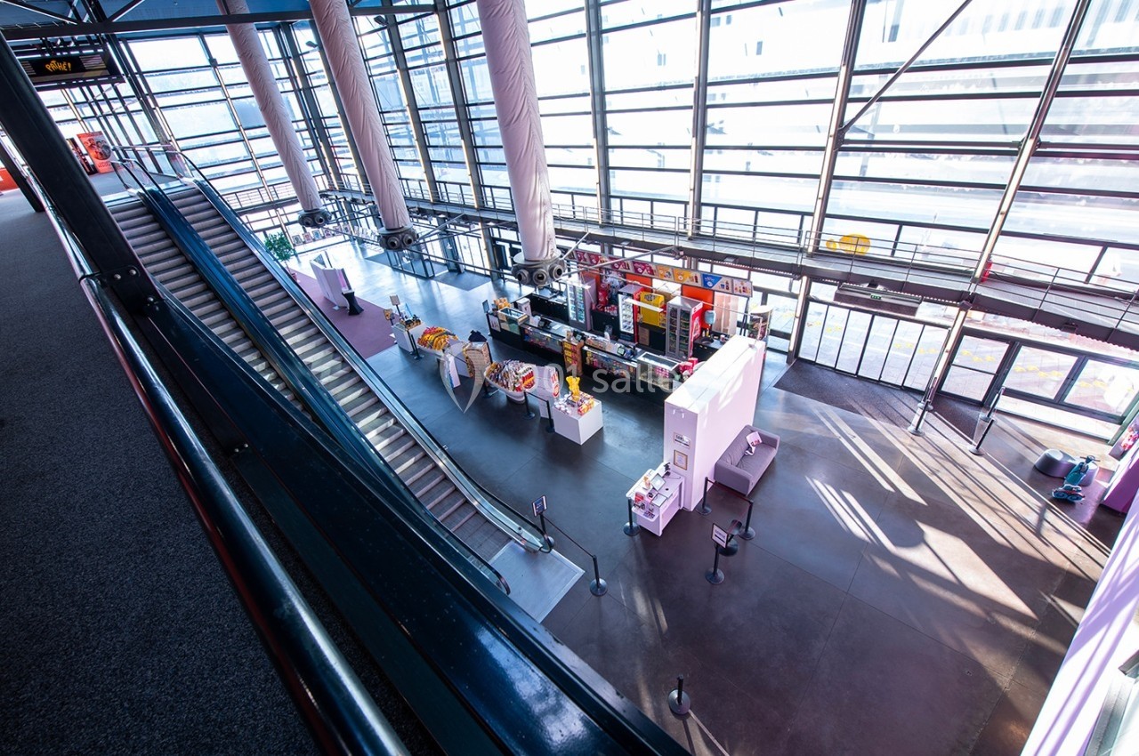 Vue intérieure d'un hall lumineux avec des stands colorés, des escaliers roulants et de grandes baies vitrées.