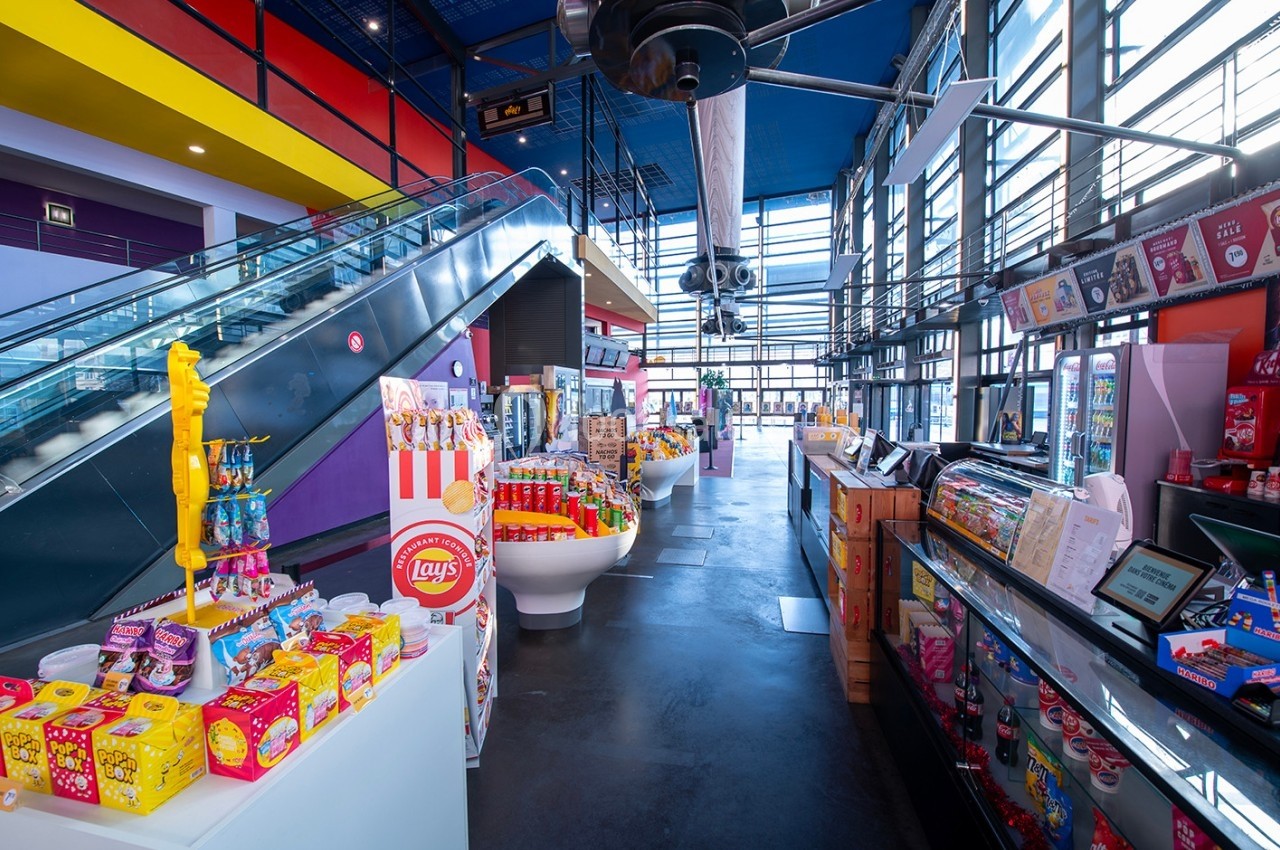 Magasin coloré avec des étagères remplies de confiseries et snacks, escalier roulant et grandes fenêtres en arrière-plan.