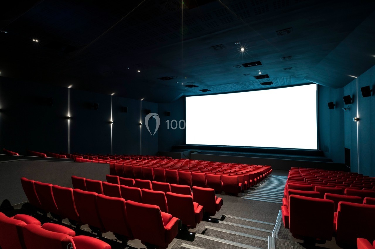 Salle de cinéma vide avec des rangées de sièges rouges et un grand écran blanc allumé.