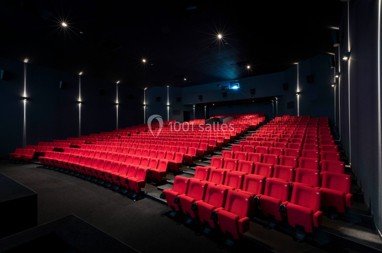 Salle de cinéma vide avec des rangées de sièges rouges et un écran au fond, éclairée par des lumières tamisées.