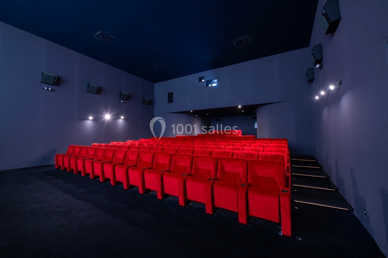 Salle de cinéma vide avec des rangées de fauteuils rouges, murs gris et éclairage tamisé.