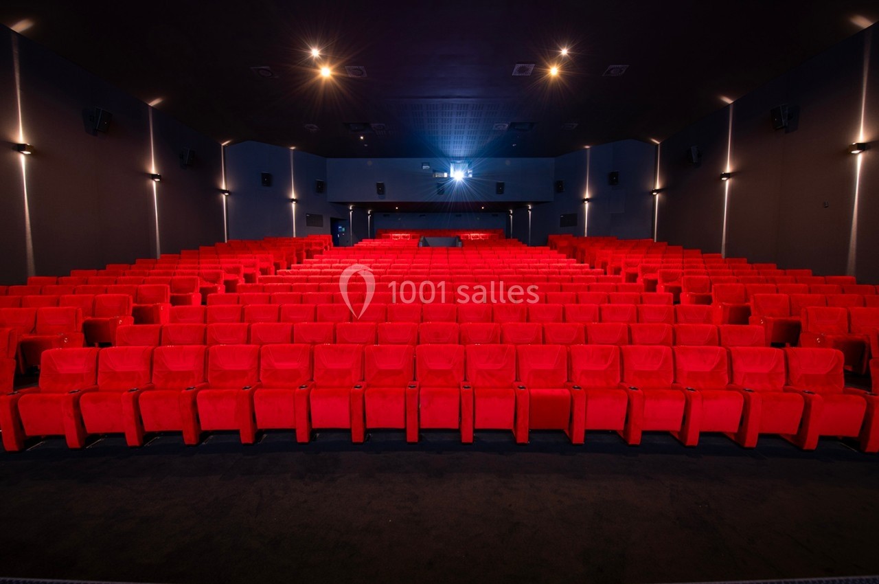 Salle de cinéma vide avec rangées de sièges rouges et écran éclairé au fond.