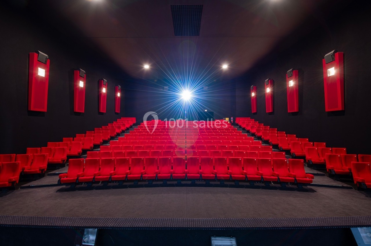 Salle de cinéma vide avec des rangées de sièges rouges et un projecteur allumé au fond.