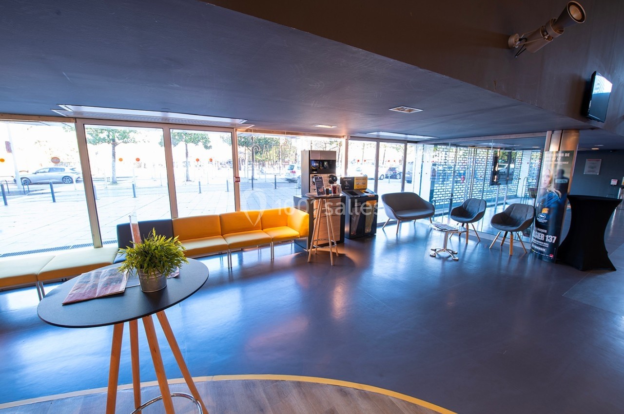 Espace d'accueil lumineux avec canapés jaunes, chaises grises, table haute et grandes baies vitrées donnant sur l'extérieur.