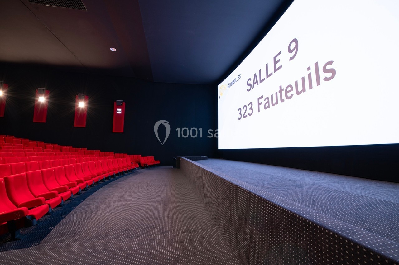 Salle de cinéma vide avec des sièges rouges, un écran affichant ’Salle 9, 323 fauteuils’ et un éclairage tamisé.