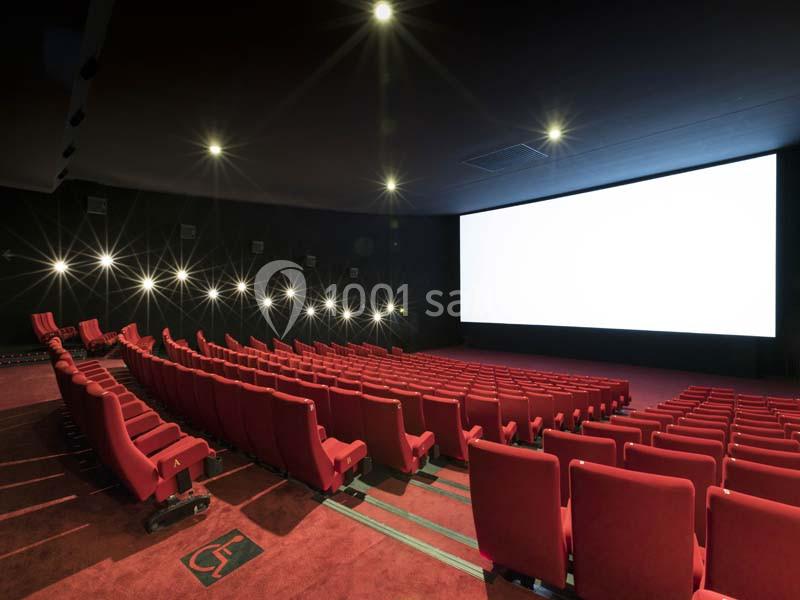 Salle de cinéma vide avec des rangées de fauteuils rouges et un grand écran blanc allumé.