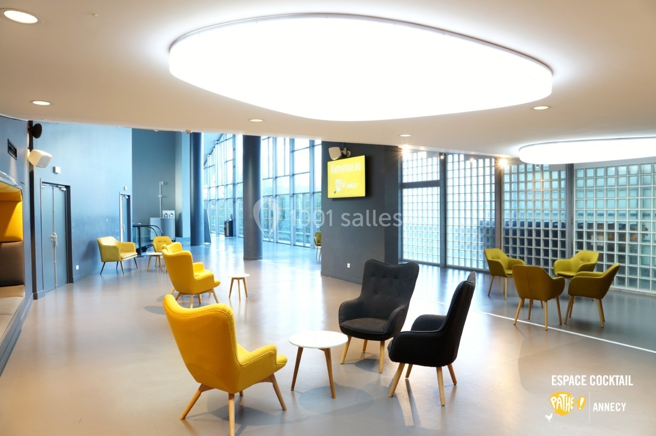 Salle lumineuse avec fauteuils colorés, tables basses et grande baie vitrée donnant sur l'extérieur.