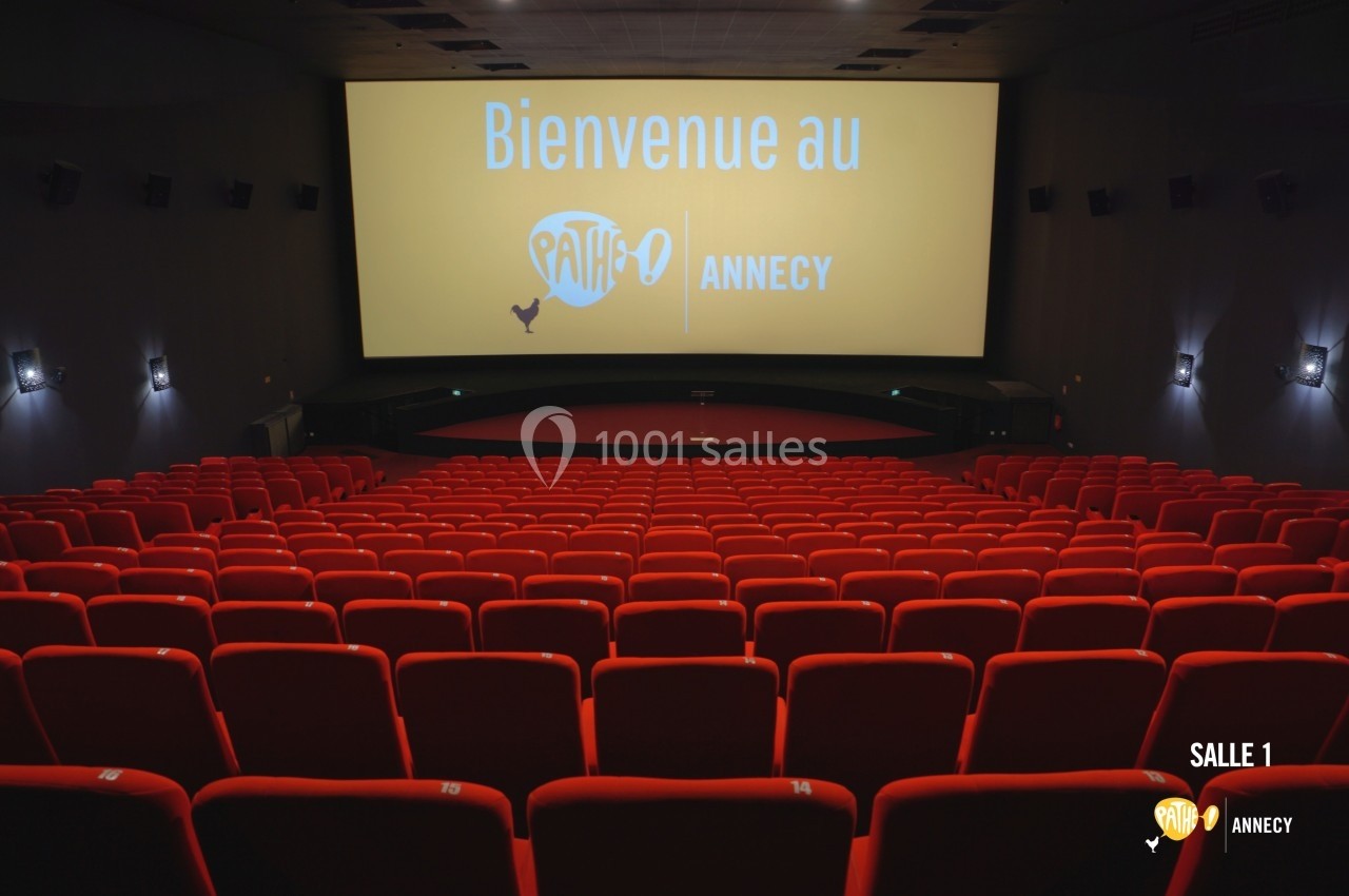 Salle de cinéma vide avec des rangées de sièges rouges et un écran affichant ’Bienvenue au Pathé Annecy’.