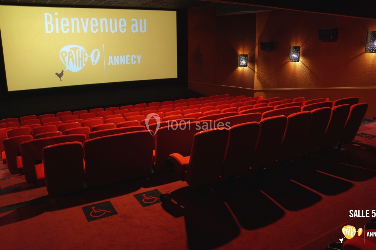 Salle de cinéma avec rangées de sièges rouges, écran affichant ’Bienvenue au Pathé Annecy’ et espaces accessibles au premier…