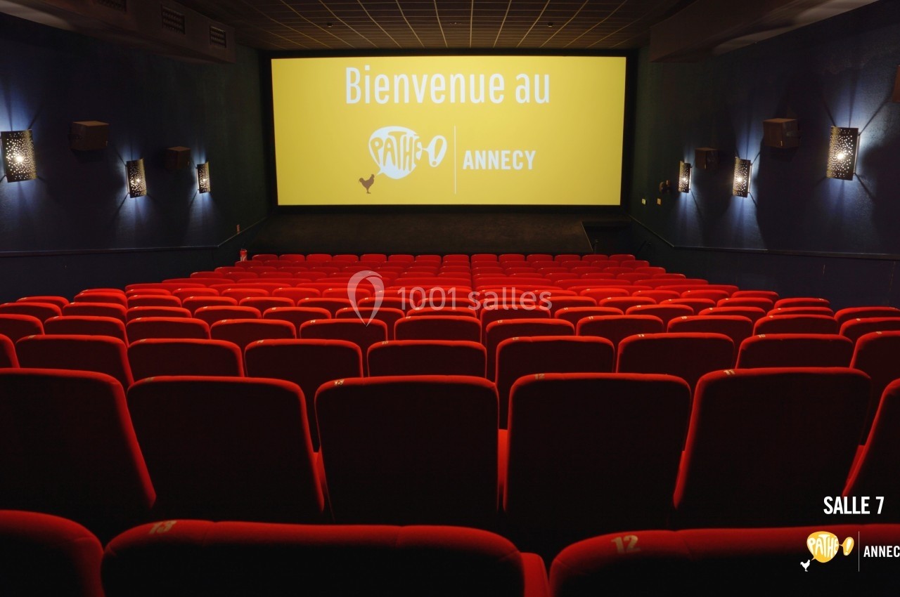 Salle de cinéma vide avec des rangées de sièges rouges et un écran affichant ’Bienvenue au Pathé Annecy’.