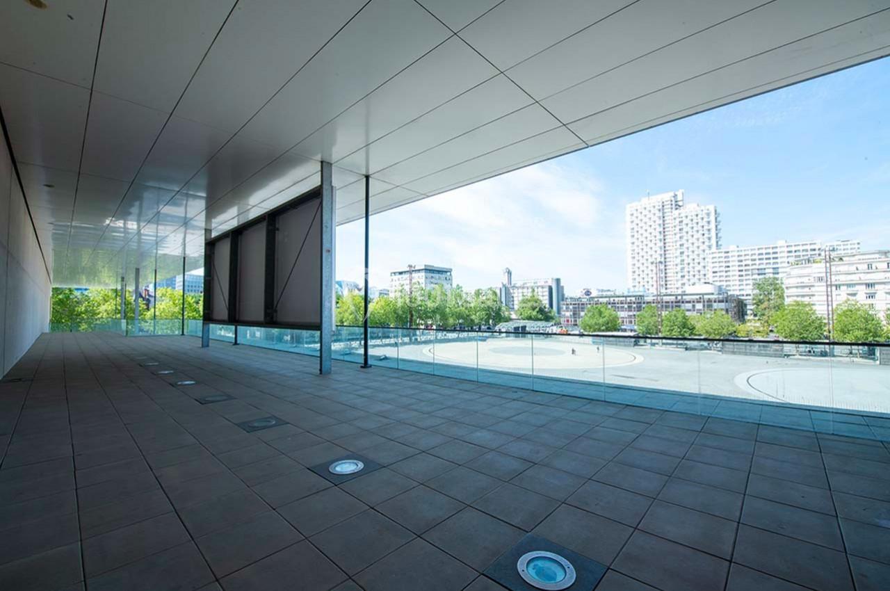 Vue d'une terrasse couverte avec sol carrelé, balustrade en verre et panorama urbain sous un ciel dégagé.