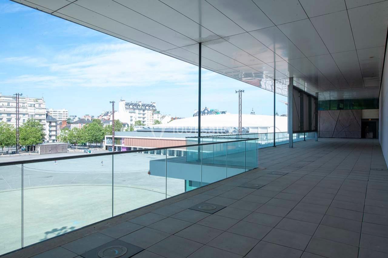 Vue depuis une terrasse couverte avec garde-corps en verre, donnant sur une place urbaine et des bâtiments modernes.