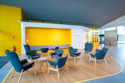 Salle lumineuse avec murs jaunes et gris, mobilier moderne espacé, grandes fenêtres laissant entrer la lumière naturelle.