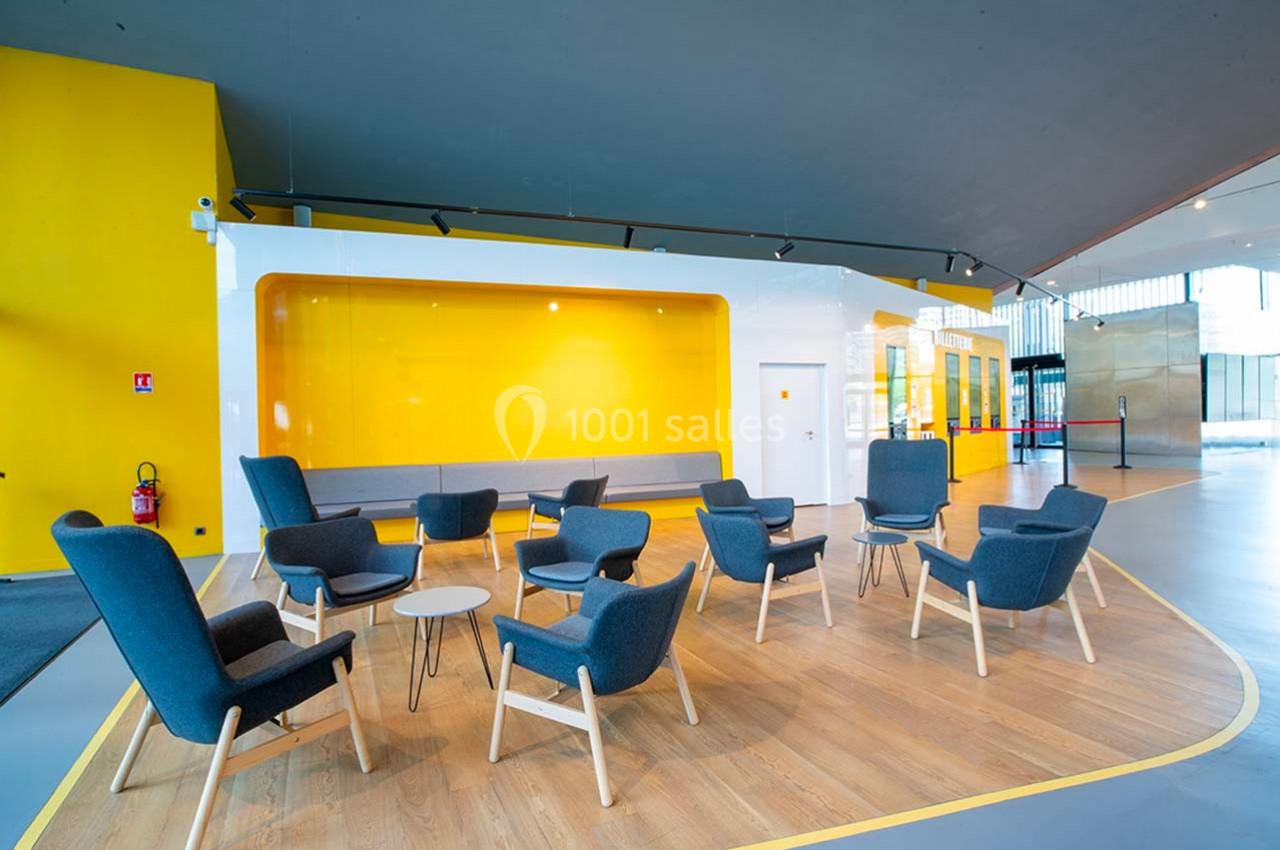 Espace d'attente moderne avec fauteuils gris, tables basses blanches et mur jaune vif en arrière-plan.