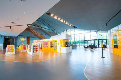 Salle lumineuse avec murs jaunes et gris, mobilier moderne espacé, grandes fenêtres laissant entrer la lumière naturelle.