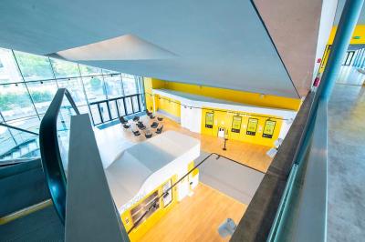 Salle lumineuse avec murs jaunes et gris, mobilier moderne espacé, grandes fenêtres laissant entrer la lumière naturelle.