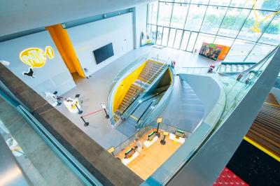 Salle lumineuse avec murs jaunes et gris, mobilier moderne espacé, grandes fenêtres laissant entrer la lumière naturelle.
