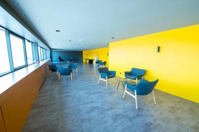 Salle lumineuse avec murs jaunes et gris, mobilier moderne espacé, grandes fenêtres laissant entrer la lumière naturelle.