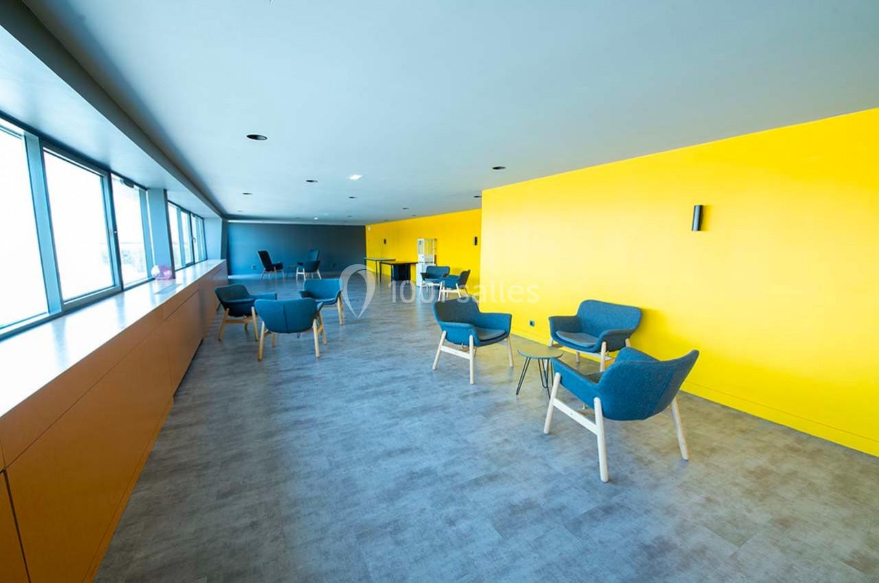 Salle lumineuse avec murs jaunes, chaises bleues, tables basses en verre et grandes fenêtres laissant entrer la lumière…