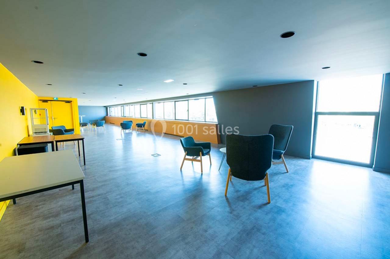 Salle lumineuse avec murs jaunes et gris, mobilier moderne espacé, grandes fenêtres laissant entrer la lumière naturelle.
