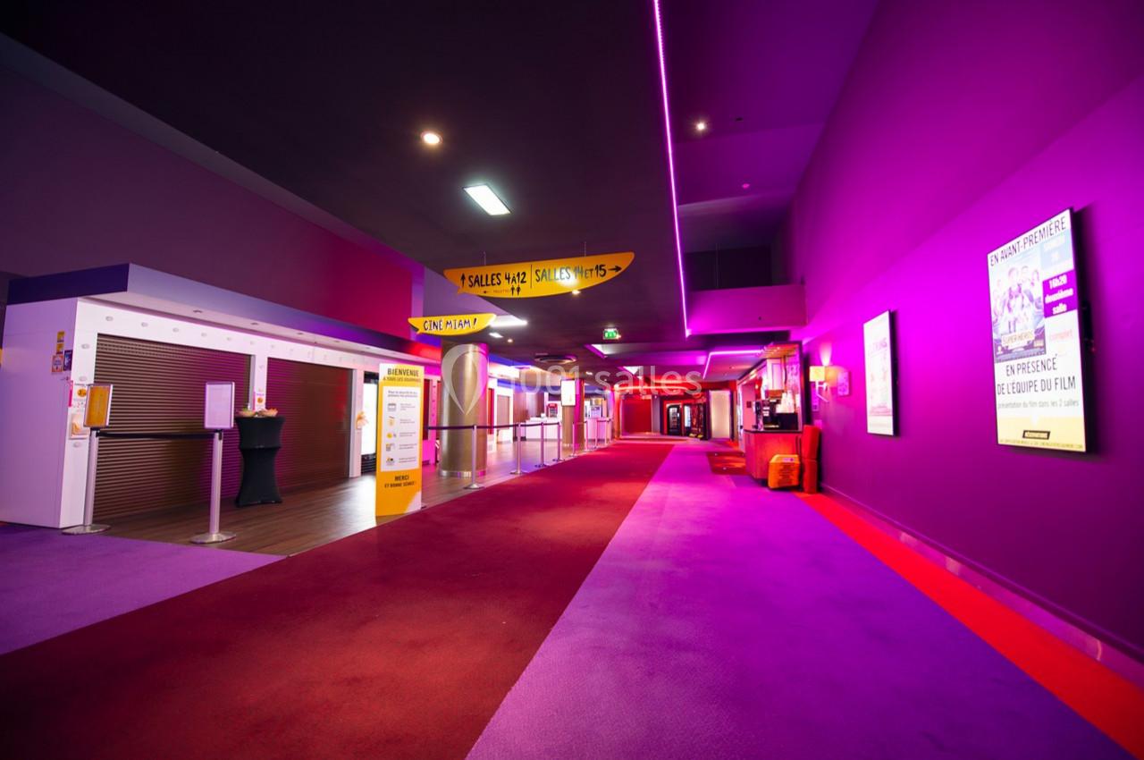 Couloir d'un cinéma avec moquette rouge et éclairage violet, affiches et panneaux d'information visibles.