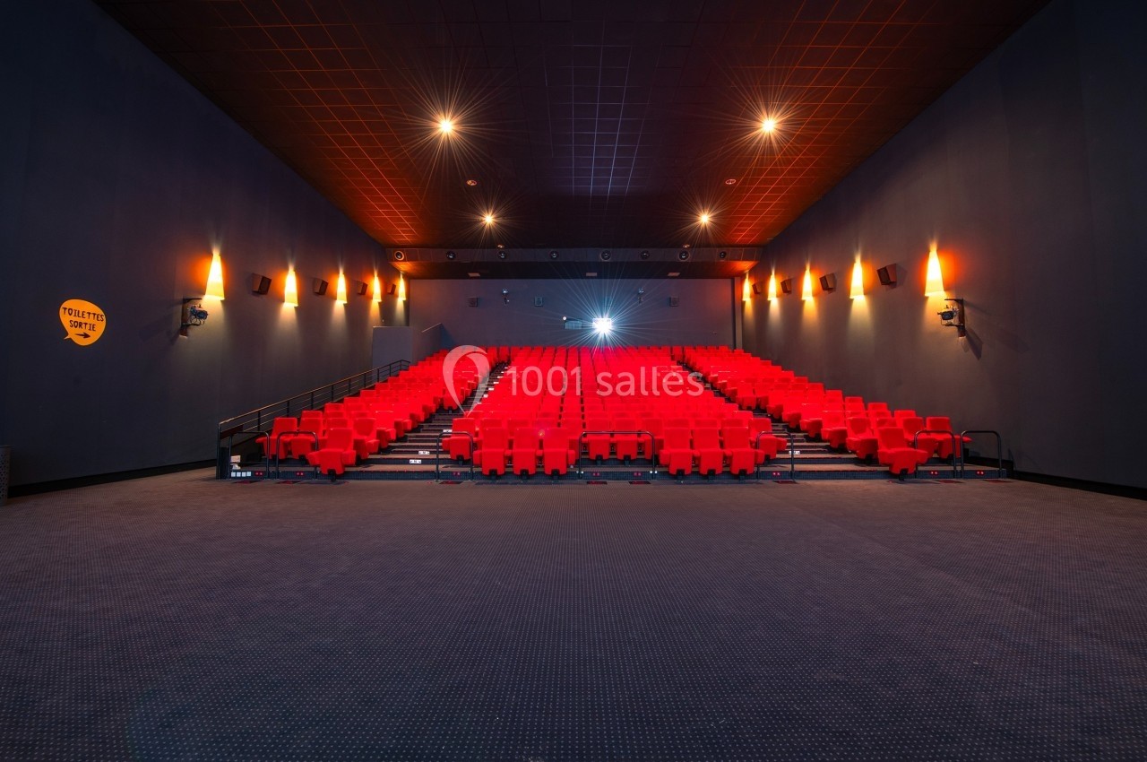Salle de cinéma vide avec rangées de sièges rouges, éclairage tamisé et écran éteint au fond.