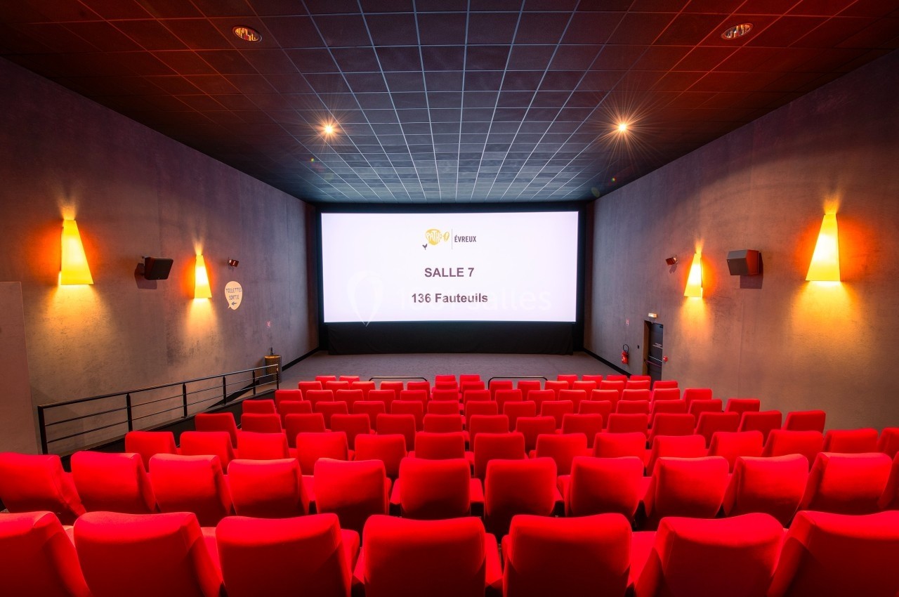 Salle de cinéma vide avec des rangées de fauteuils rouges, écran blanc affichant ’Salle 7, 136 fauteuils’.