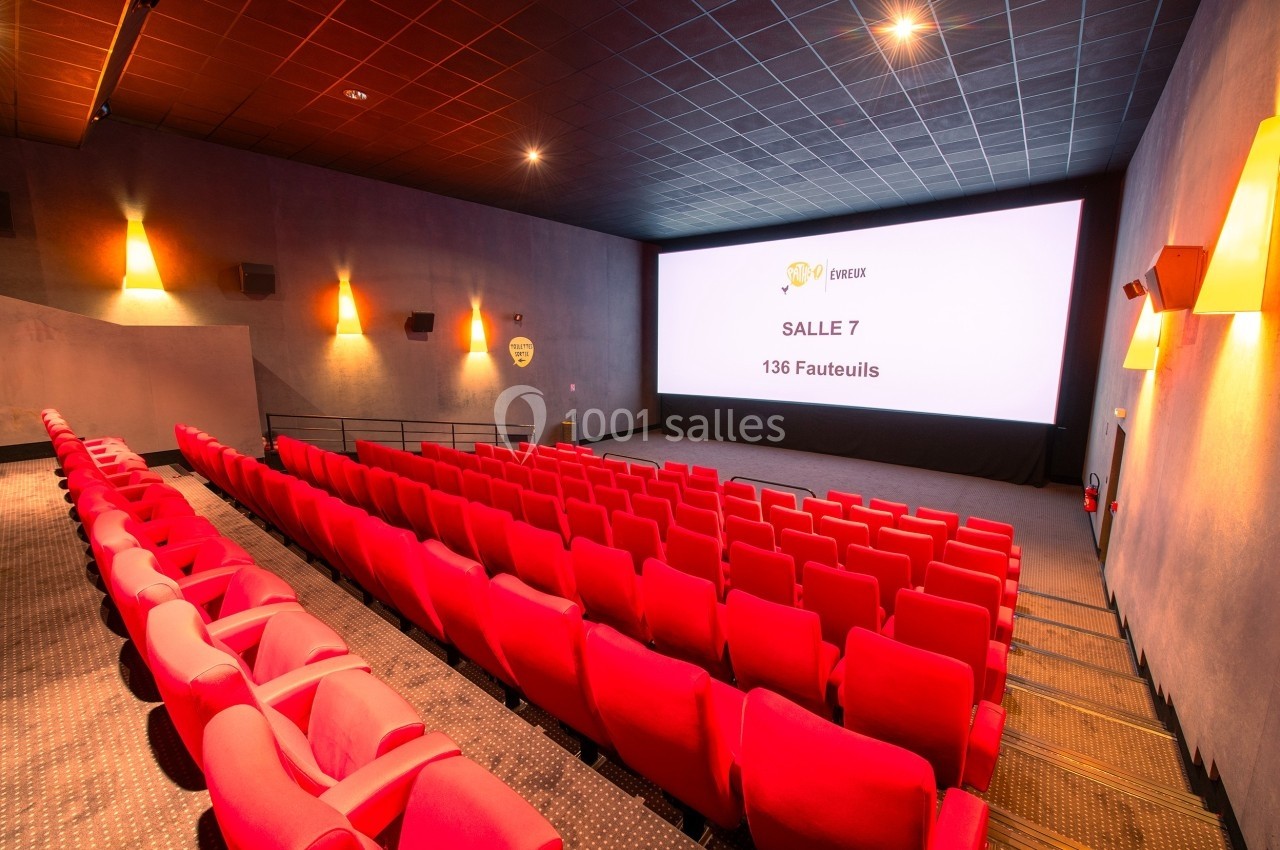 Salle de cinéma vide avec rangées de fauteuils rouges, écran blanc affichant ’Salle 7 - 136 fauteuils’.