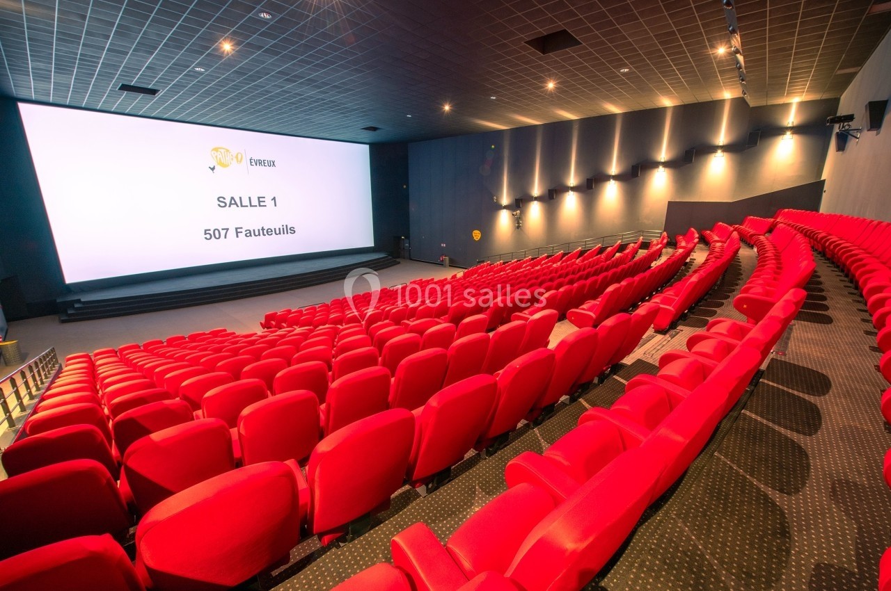 Salle de cinéma moderne avec rangées de fauteuils rouges vides, grand écran et éclairage tamisé.