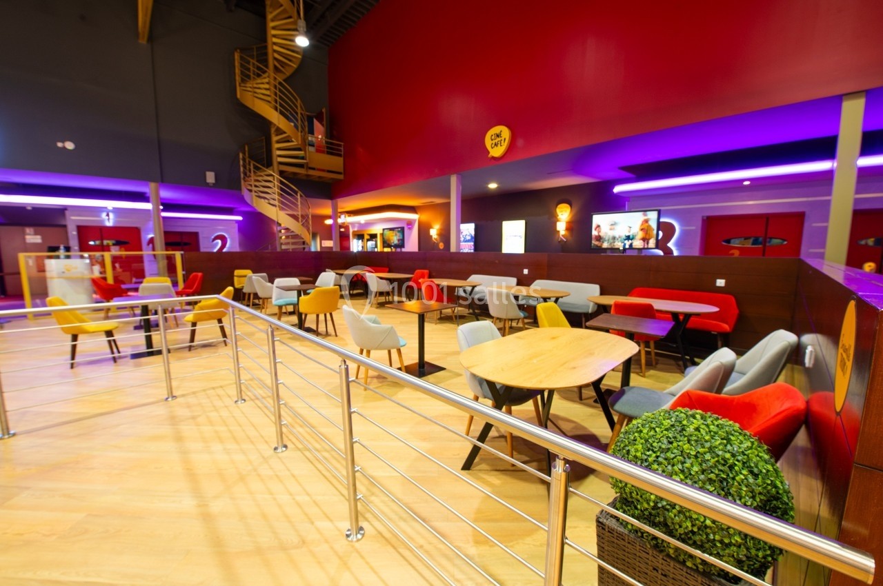 Salle lumineuse avec tables et chaises colorées, escalier en colimaçon et éclairage violet au plafond.