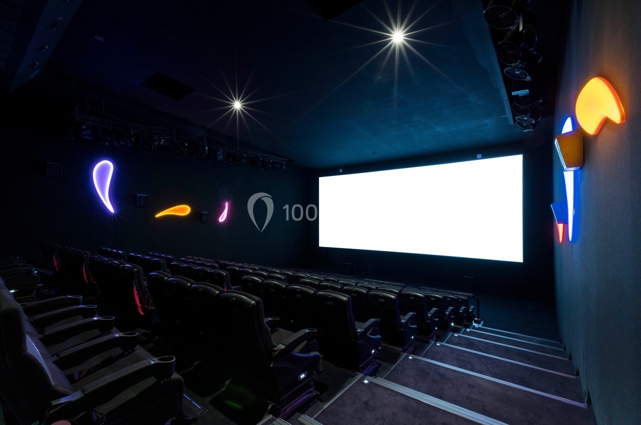 Salle de cinéma sombre avec des rangées de sièges vides, un écran blanc allumé et des lumières néon colorées sur les murs.