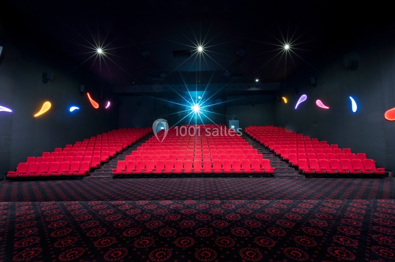 Salle de cinéma vide avec des rangées de sièges rouges, un projecteur allumé et un sol à motifs sombres.