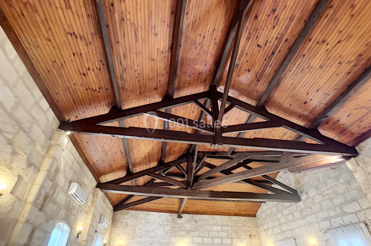 Plafond en bois avec poutres apparentes dans une salle aux murs en pierre, éclairée par des appliques murales.