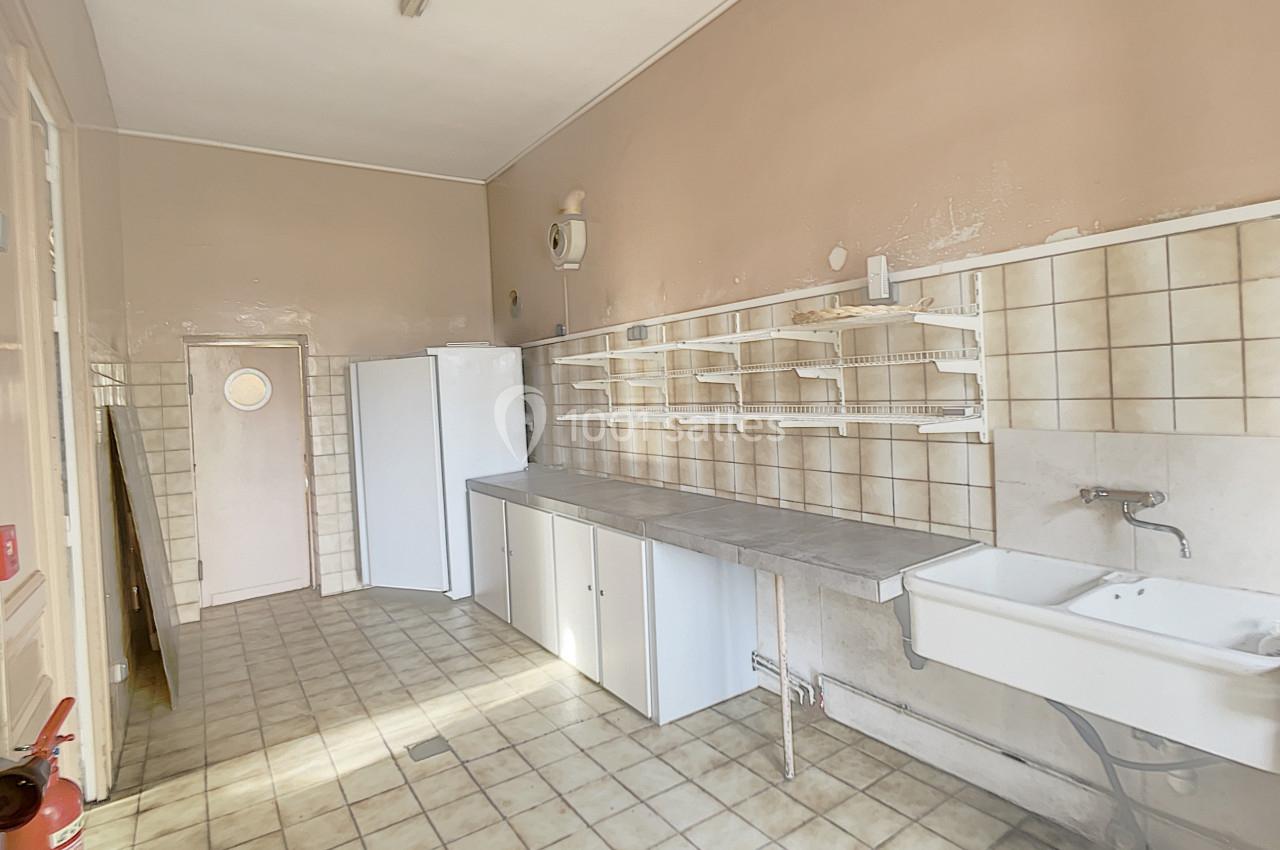 Cuisine vide avec carrelage beige, évier mural, étagères blanches, réfrigérateur et porte au fond.