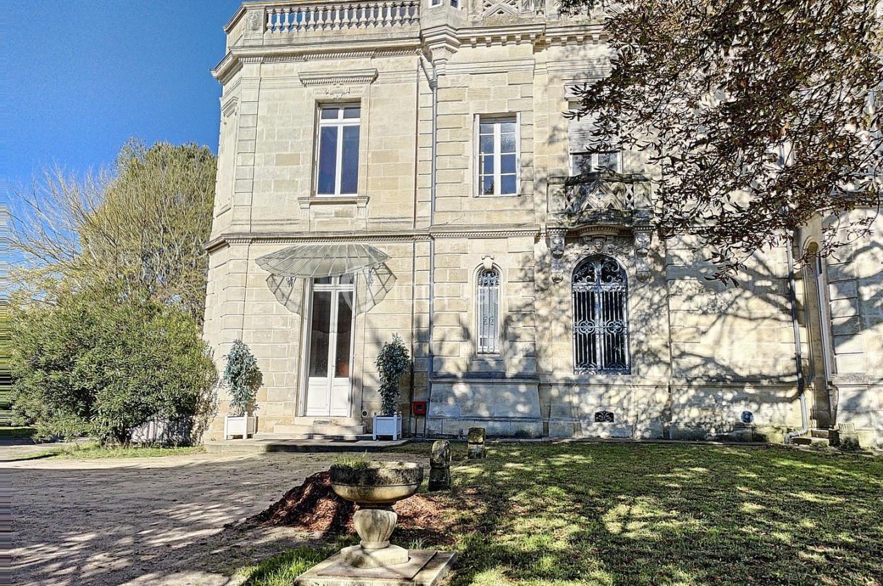 Façade d'un bâtiment en pierre avec fenêtres ornées, entouré d'arbres et d'une pelouse ensoleillée.